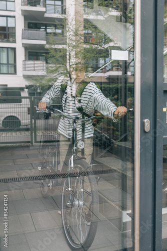 A teenage is riding a bicycle in front of a building, the reflection of the person and the bicycle can be seen in the window