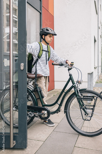 A teenager girl wearing a helmet and a backpack is standing next to a bicycle leaving your home