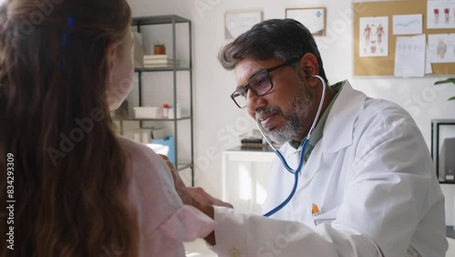 Medium close-up of Indian male pediatrician examining young Caucasian girl sitting on couch, listening to chest with stethoscope, asking to breathe in and out, checking eyes, during follow-up visit