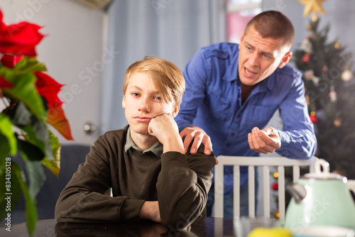 Portrait of upset young schoolboy siiting at table and quarreling with his father during xmas weekand photo