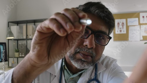 Medium close-up POV shot of middle-aged Asian male physician examining patients eye, shining torch through pupil, studying fundus and blood vessels, while testing eyesight in clinic