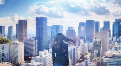 日本の街・都市・朝日を受けたオフィスビル群を前に佇み風を受けて髪をなびかせる会社員の女性の出勤　就職・ビジネス・はじまり・決意・目標・挑戦のイメージ © tenpadasi