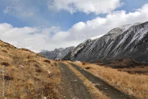 A field road climbs a grassy hill after rain to meet snow-covered mountain ranges. © Алексей Желтухин