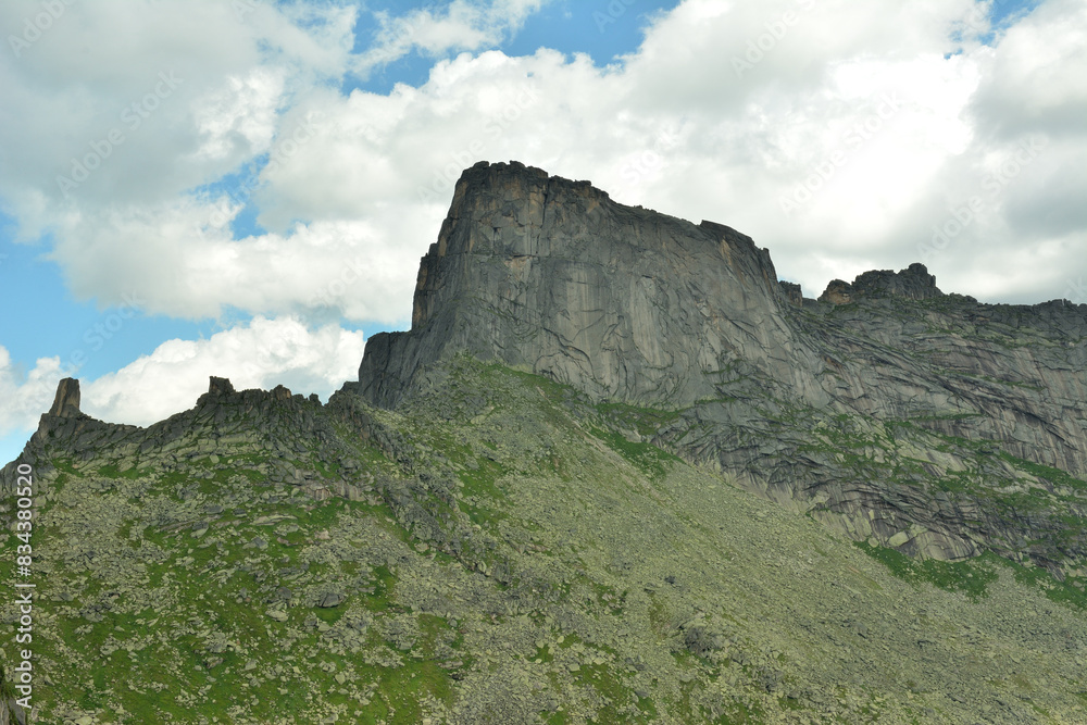 A tall rock with pointed peaks and steep slopes against the backdrop of a summer cloudy sky.