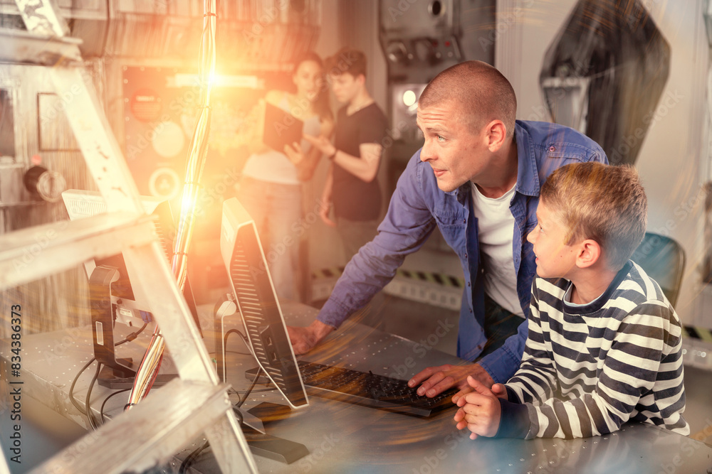 Fototapeta premium Interested young man with his tween son using computer to find solution to puzzle in escape room stylized as abandoned bunker. Toned image with visual effects