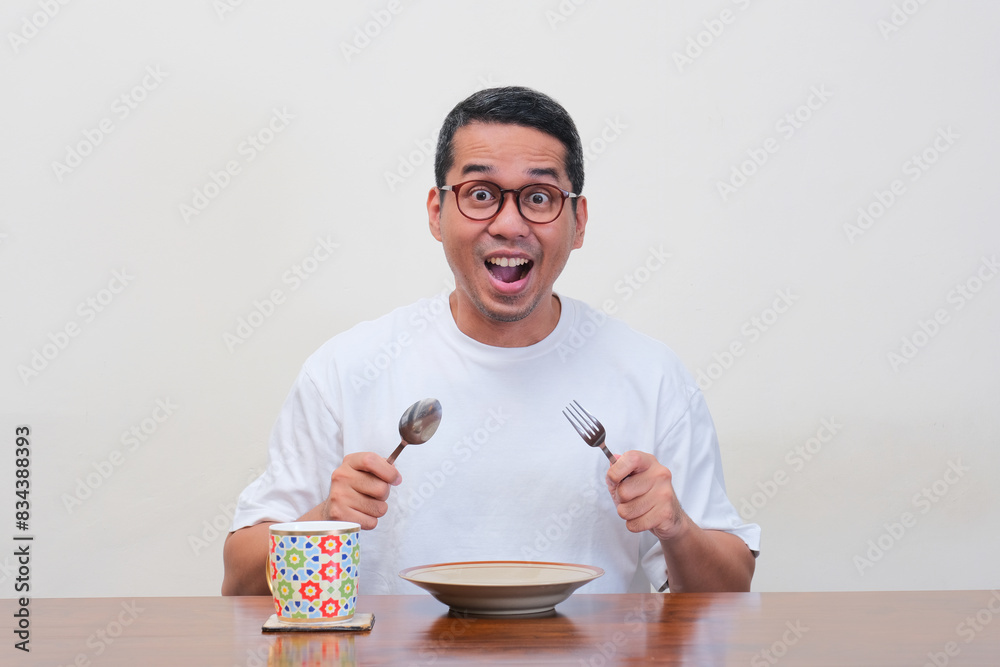 A man sitting on dinning table holding spoon and fork showing wow face expression A man sitting on dinning table holding spoon and fork showing wow face expression