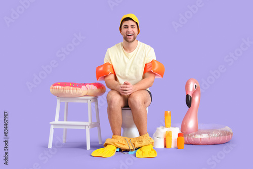 Male vacationer with beach accessories sitting on toilet bowl against lilac background photo