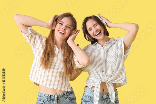 Female friends in headphones listening to music on yellow background