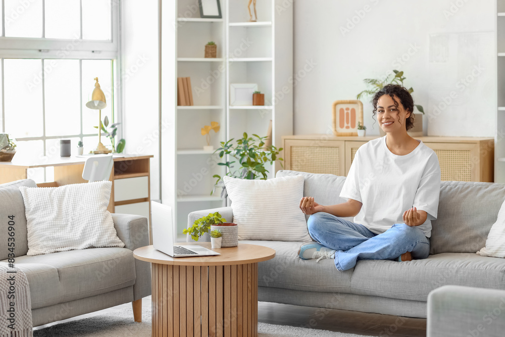 Young African-American woman meditating on sofa with laptop at home Young African-American woman meditating on sofa with laptop at home