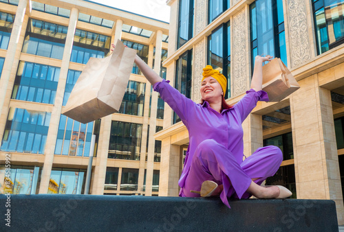 Obraz na płótnie woman with craft shopping bags in the city. The concept of fashionable Consumerism and ecology. An adult Asian woman in a purple suit with red and yellow accents enjoys shopping . a sunny day. w salonie