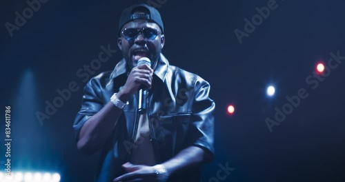 RnB Musician Gives a Breathtaking Performance Under a Canopy of Lights. Stunning Visual Effects Enrich the Hip Hop Concert. Zoom In Portrait of a Black Man in Leather Jacket, Shades and a Cap photo