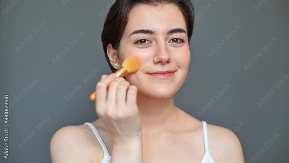 Women's cosmetics beauty, young smiling woman retouching skin with powder looking at camera standing on gray background Women's cosmetics beauty, young smiling woman retouching skin with powder looking at camera standing on gray background