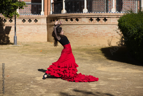 Beautiful woman dancing flamenco in Seville, Spain. She is wearing a red and black gypsy dress and dancing flamenco with a lot of art. In the background a monument with arches and columns.