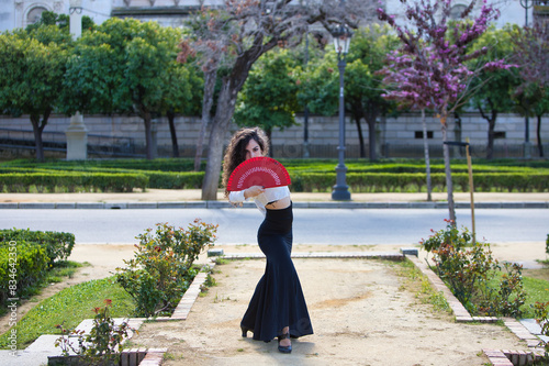 Beautiful woman with long curly hair, dancing flamenco with a red fan, she is in Seville, Spain. She wears long black skirt, white shirt and makes different dance poses. Flamenco, heritage of humanity
