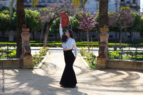Beautiful woman with long curly hair, dancing flamenco with a red fan, she is in Seville, Spain. She wears long black skirt, white shirt and makes different dance poses. Flamenco, heritage of humanity