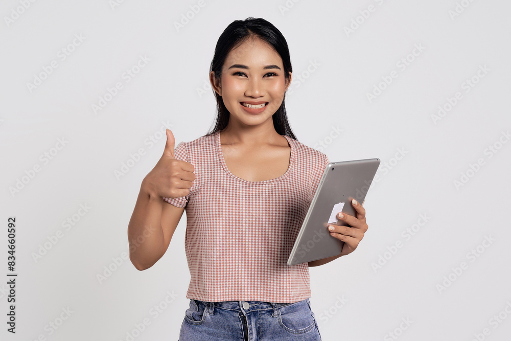 Smiling cheerful happy beautiful young Asian woman in a casual t-shirt holding digital tablet pc computer and showing thumb up isolated on white background Smiling cheerful happy beautiful young Asian woman in a casual t-shirt holding digital tablet pc computer and showing thumb up isolated on white background