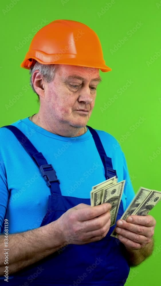 Senior construction worker in overalls counting dollars and puts it in pocket. Senior construction worker in overalls counting dollars and puts it in pocket.