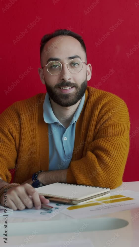 Medium vertical portrait of Middle Eastern male creative communications agency executive or director sitting in office with business documents on table, against red wall, smiling for camera