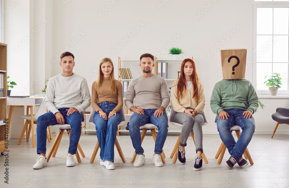 Naklejka premium Confident smiling people job candidates and seekers wearing casual clothes sitting in a row with unknown faceless man in paper bag on head with question mark. Employment and recruitment concept