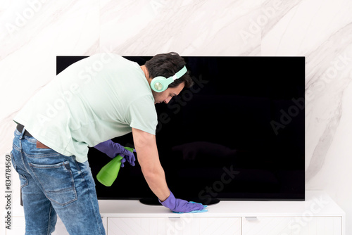 Young man in headphones wiping television with a rag and spray