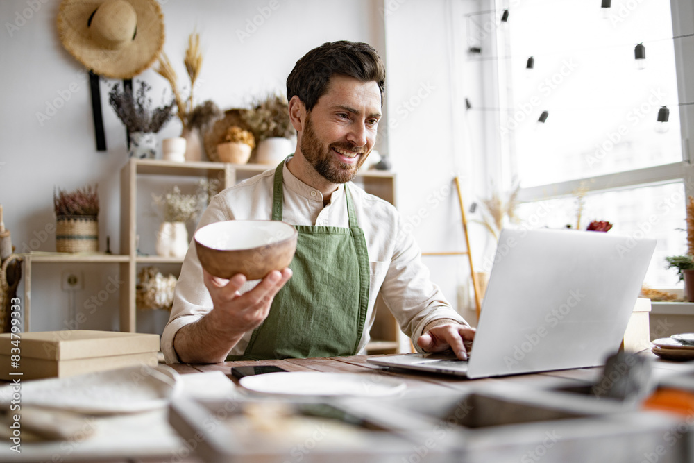 Adult bearded male potter engaged in e-commerce selling handmade clay products on Internet. Happy worker in apron holds unique bowl created in personal style against background of shelves with dishes. Adult bearded male potter engaged in e-commerce selling handmade clay products on Internet. Happy worker in apron holds unique bowl created in personal style against background of shelves with dishes.