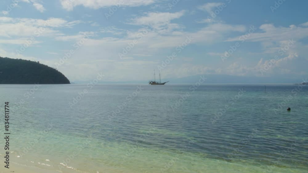 A distant sailboat rests anchored off Kri Island in Raja Ampat, Indonesia. The clear, inviting waters and vast, open sky evoke a sense of peace and adventure. Static slow motion shot. A distant sailboat rests anchored off Kri Island in Raja Ampat, Indonesia. The clear, inviting waters and vast, open sky evoke a sense of peace and adventure. Static slow motion shot.