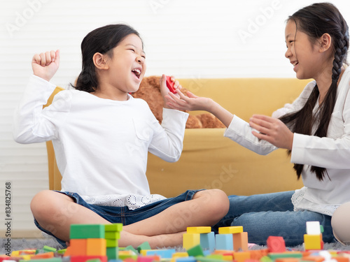 Adorable Asian kid playing wooden blocks with her sister on the floor of living room. Happy sibling children have fun playing game toy together at home. with colorful blocks. Playful girls enjoyment.