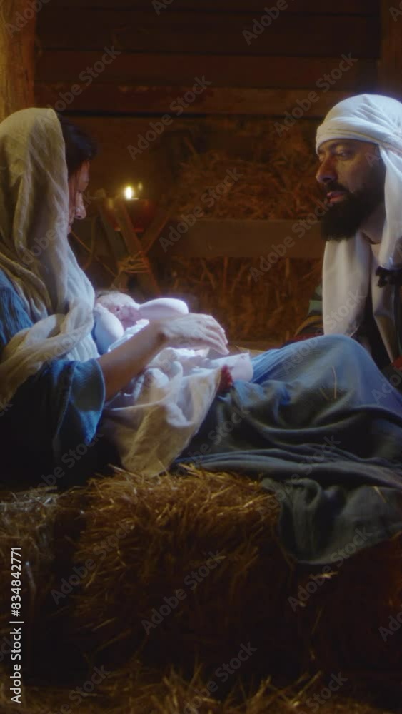 Zoom in view of Joseph talking with Mother Mary caressing baby Jesus on Christmas day in dark stable in Bethlehem. Vertical shot. Zoom in view of Joseph talking with Mother Mary caressing baby Jesus on Christmas day in dark stable in Bethlehem. Vertical shot.