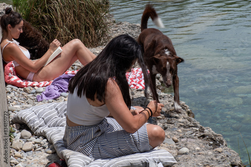 Woman chatting on mobile phone in riverscape, dressed in striped trousers and white tank top, long black hair, in the background legs of a girl sunbathing and reading, her dog walking next to her.