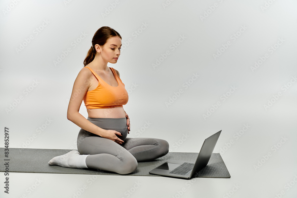 Naklejka premium A sporty pregnant woman sits on a yoga mat, multitasking with a laptop.
