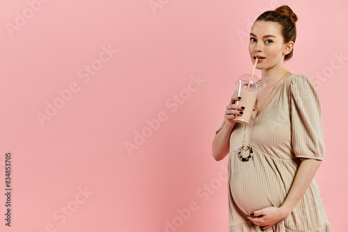 A young pregnant woman in a dress holding milkshake on a pink background.