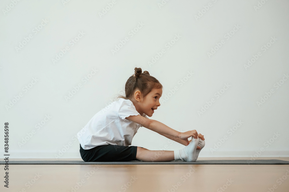 Side view, stretching. Cute little girl is sitting on the yoga mat and doing exercises