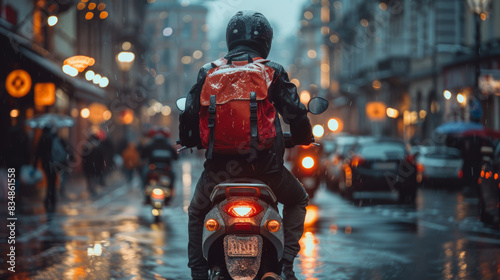 Captured from behind, a motorcyclist wearing a helmet and red backpack rides through a rain-soaked city street at night with glowing lights © AS Photo Family