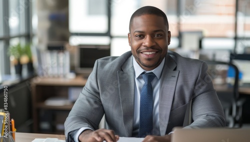 A handsome African American businessman in his late thirties, wearing grey suit and blue tie is sitting at the desk smiling while looking into camera.