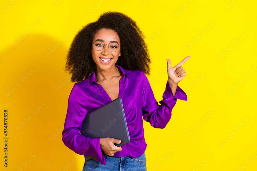 Photo portrait of lovely young lady point empty space hold netbook dressed stylish violet garment isolated on yellow color background Photo portrait of lovely young lady point empty space hold netbook dressed stylish violet garment isolated on yellow color background