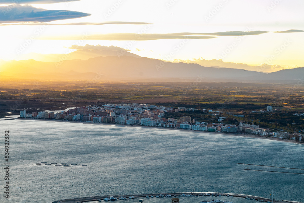La fotografía muestra una vista panorámica de una ciudad costera al atardecer, iluminada por la luz dorada del sol poniente. Se observan montañas en el horizonte y el mar en primer plano. La fotografía muestra una vista panorámica de una ciudad costera al atardecer, iluminada por la luz dorada del sol poniente. Se observan montañas en el horizonte y el mar en primer plano.