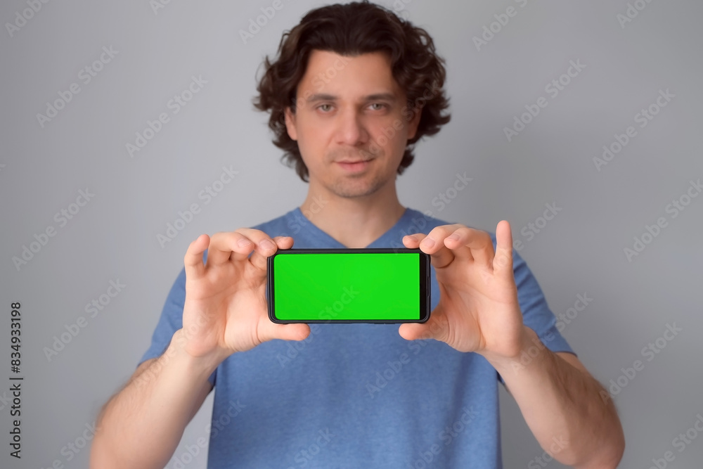 Man holding in hands smartphone with green screen on gray background. Curly brunet guy wearing blue t-shirt looking at camera showing mobile phone in focus. Advertising banner poster, advertisement. Man holding in hands smartphone with green screen on gray background. Curly brunet guy wearing blue t-shirt looking at camera showing mobile phone in focus. Advertising banner poster, advertisement.