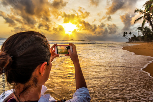 Young woman taking photo of beautiful tropical beach on smartphone at sunset