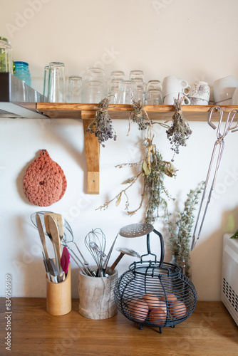 Rustic kitchen detail, bowl with spoons and ladles, shelf with glasses and cups. Hanging aromatic herbs and eggs in container.