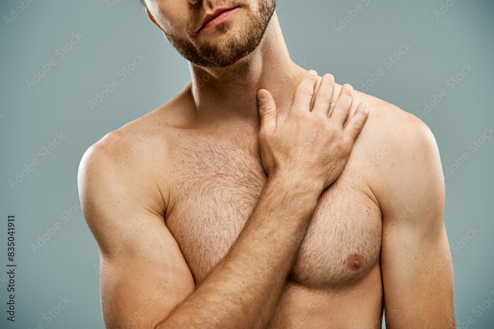 A rugged, shirtless man with a beard confidently stands with hands on his chest on a grey studio backdrop. A rugged, shirtless man with a beard confidently stands with hands on his chest on a grey studio backdrop.