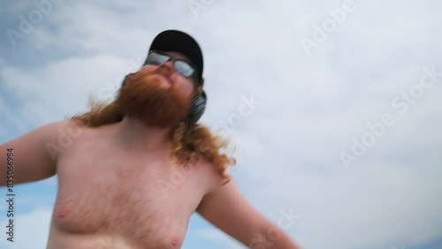Bearded hipster relaxed man love drinking beer dancing on summer vacation looking at camera with phone in hand. Low angle.
