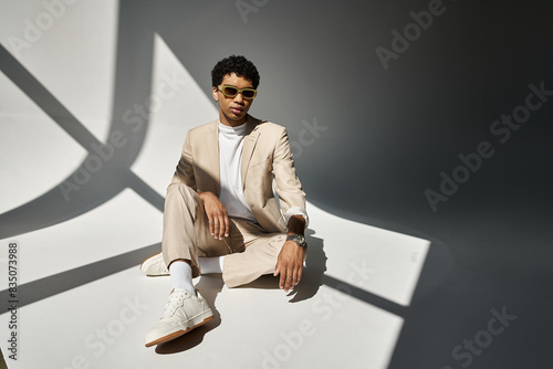 Tableau sur toile African American man in beige suit sitting comfortably on the floor, wearing stylish sunglasses