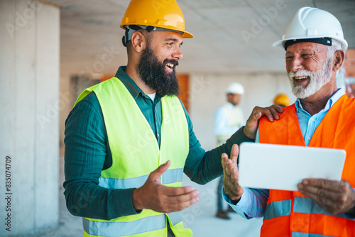 A diverse duo, a construction worker in a yellow helmet and a site manager wearing a white helmet, review plans on a tablet at a construction site, engaged in a cheerful conversation.