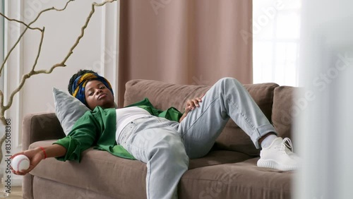 African American girl lies on couch in room and throws up baseball up and down, feeling lonely or bored. Black young woman is waiting for someone call or text, lost in thought. melancholy and boredom photo