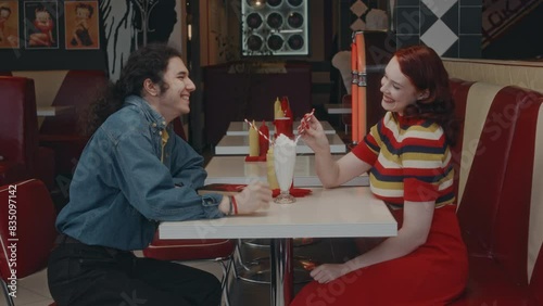 Side medium long shot of smiling Caucasian boyfriend and girlfriend drinking same milkshake in 1980s style diner, holding hands and flirting while on date