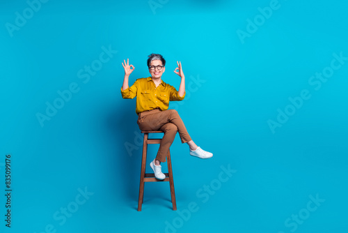 Full length photo of confident attractive lady dressed shirt glasses sitting stool showing okey signs emtpy space isolated blue color background photo