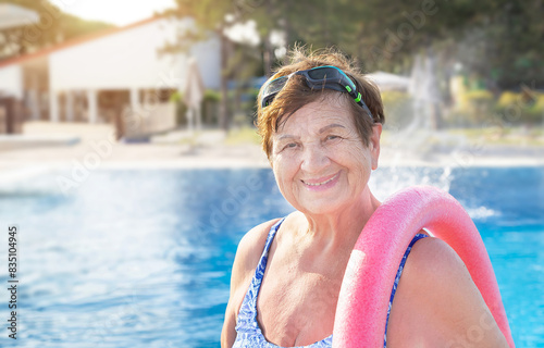 Active senior (elderly) woman (over age of 50) in sport goggles, swimsuit and with swim noodles near swimming pool smiling with happy face after swimming in summer day.