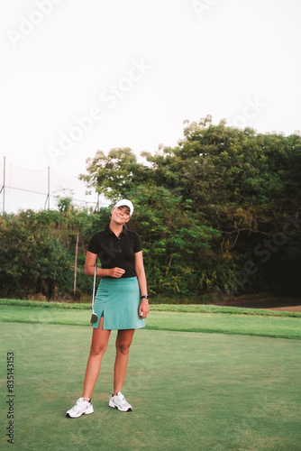 A young attractive woman who plays golf professionally on a green lawn. She is holding a golf club in her hands and looking at the camera smiling. Luxurious summer sport.