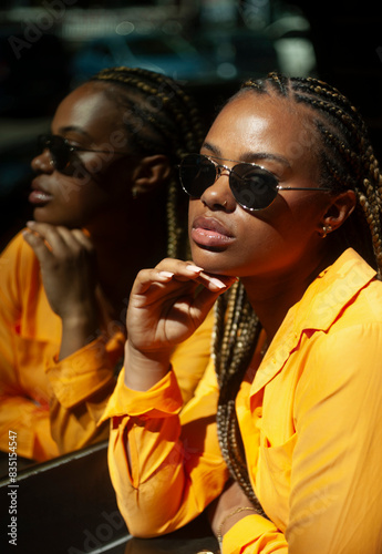 Young woman in an orange shirt and braided hair stands before a reflective surface