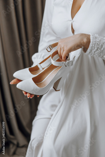 A woman is holding a pair of white shoes. The shoes are high heels and are likely for a special occasion, such as a wedding. The woman is dressed in a white dress, which complements the shoes